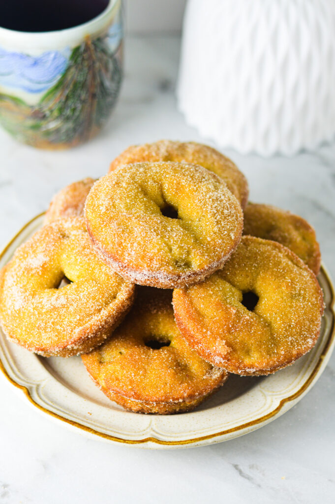 A ceramic plate with Cinnamon Sugar Banana Bread Donut arranged in a circular pattern. The background shows a cup of coffee and a white flower vase.