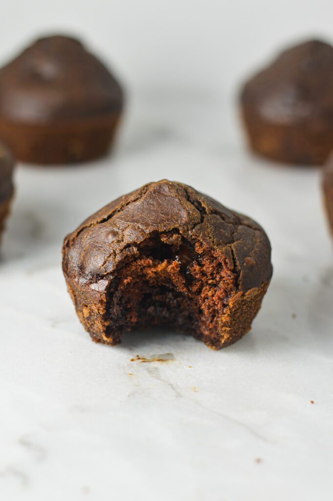 Some Healthy Mocha Muffins arranged on a granite surface with a bite taken out of one revealing a moist chocolatey interior.