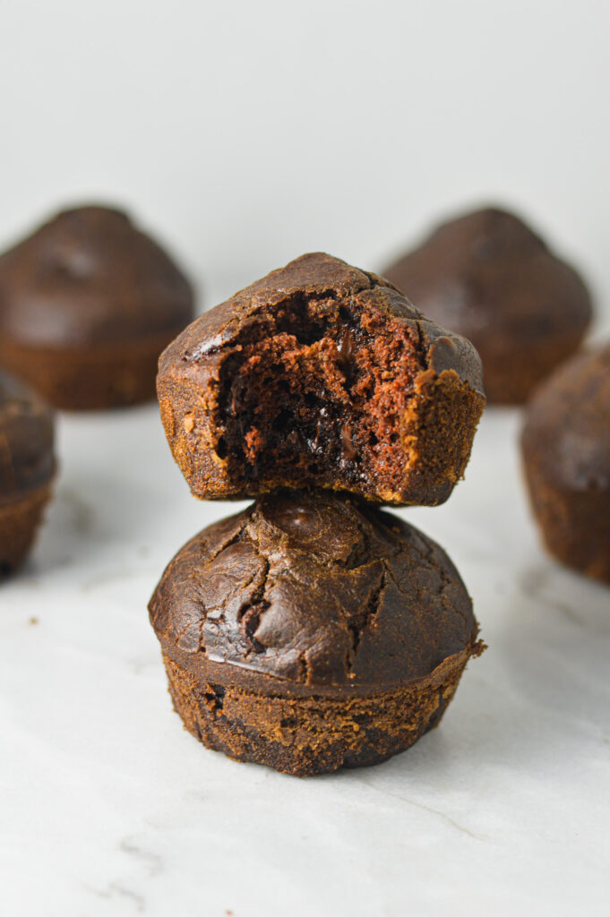 Two Healthy Mocha Muffins stacked on a white surface, with a bite taken out of the top muffin, revealing an abundance of melted chocolate chips.