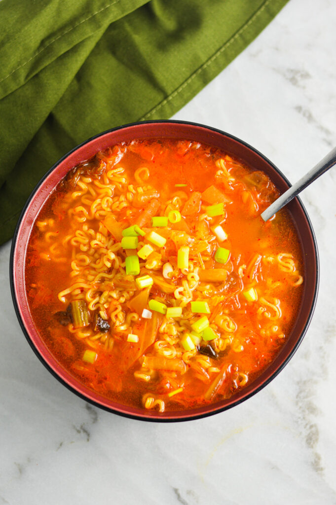 A bowl of Kimchi Ramen with curly noodles swimming in the red vibrant broth. The soup is topped with green onions.