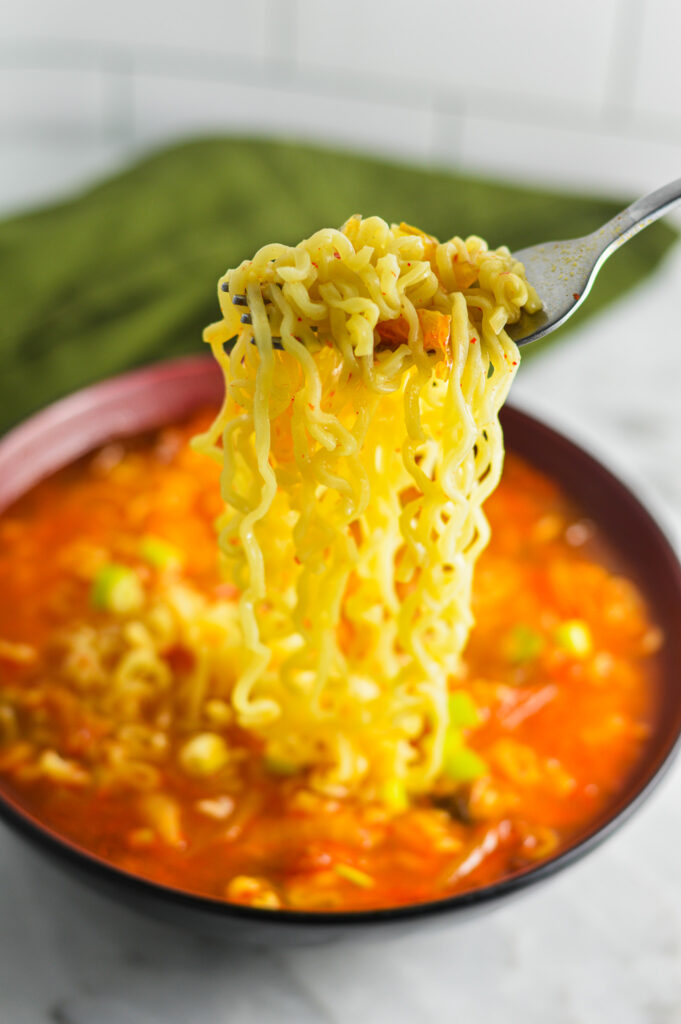 A fork pulling out some Kimchi Ramen noodles from a garnished with green onions.