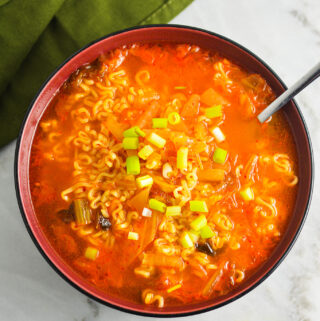 A spoon inside a red hot bowl of Kimchi Ramen, with the noodles prominently displayed in the spicy soup.