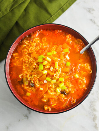 A spoon inside a red hot bowl of Kimchi Ramen, with the noodles prominently displayed in the spicy soup.