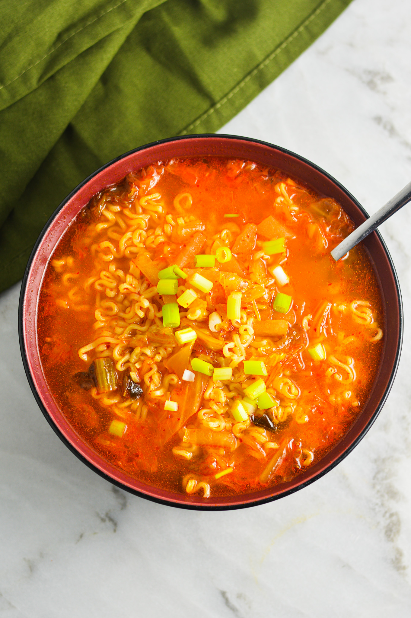 A spoon inside a red hot bowl of Kimchi Ramen, with the noodles prominently displayed in the spicy soup.