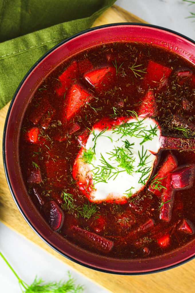 A bowl of Instant Pot Borscht on a wooden cutting board with creamy sour cream and fresh sprigs of dill on top.