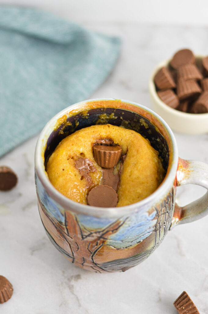 A large coffee mug with soft Peanut Butter Cup Mug Cake, and mini Reese's cups in a small bowl in the background.