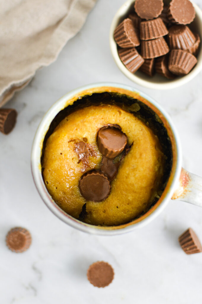 A large mug filled with Peanut Butter Cup Mug Cake, with gooey melty chocolate from the mini peanut butter cups on top of the fluffy personal sized cake.