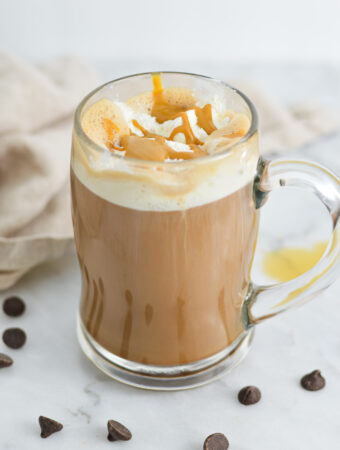 A mug filled with Peanut Butter Mocha topped with whipped cream and peanut butter drizzle, surrounded by semi-sweet chocolate chips.