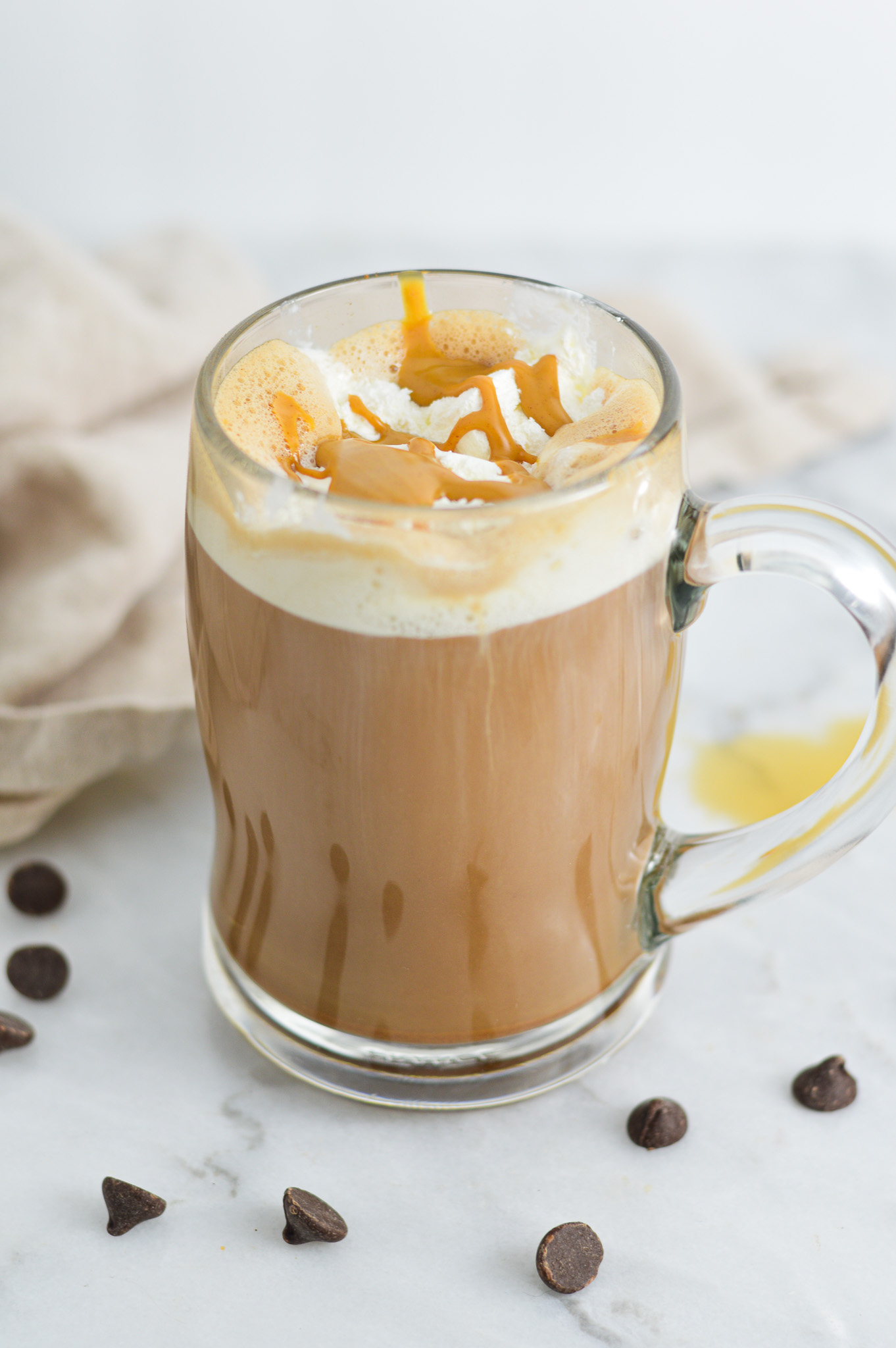 A mug filled with Peanut Butter Mocha topped with whipped cream and peanut butter drizzle, surrounded by semi-sweet chocolate chips.