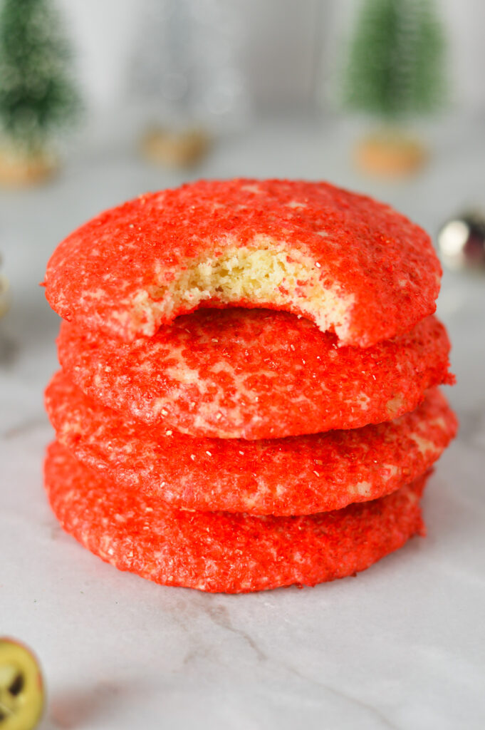 A stack of bright red Peppermint Sprinkle Cookies, with a bite taken out of the top cookie showing the fluffy texture of each cookie.