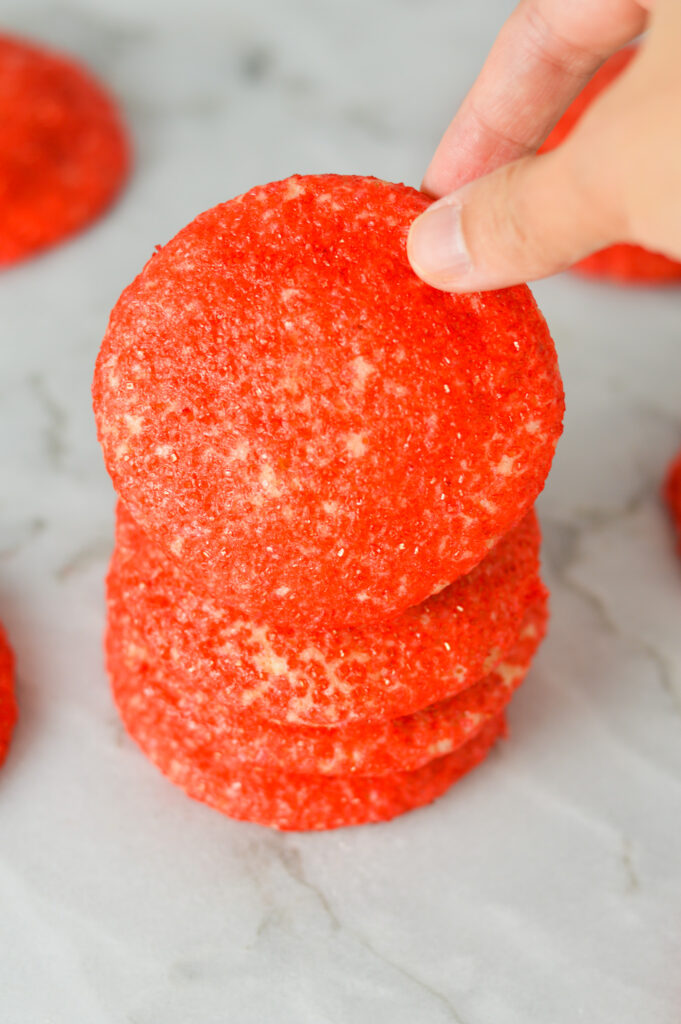 A hand picking up a Peppermint Sprinkle Cookie, showing the crunchy texture the red sparkling sugar sprinkles give the festive cookies.