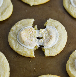 White Hot Chocolate Cookies on a cookie sheet, with a cookie cut in half showing the melted marshmallow topping.