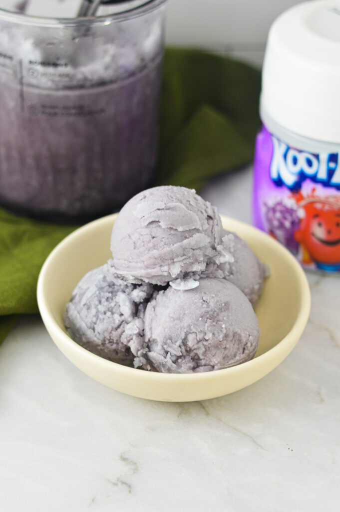 A white bowl filled with grape flavored Ninja Creami Kool-Aid Italian Ice, with a Ninja Creami pint and a cannister of kool-aid in the background.