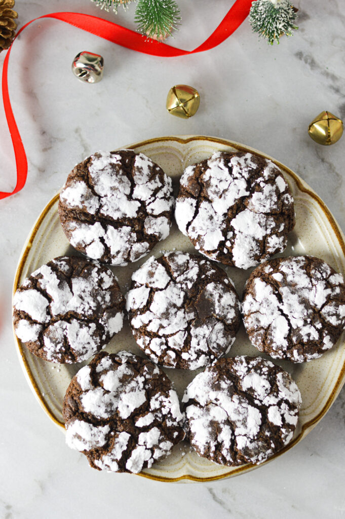 A plate of Peppermint Chocolate Crinkle Cookies with festive decor surrounding it, like bells and sprigs of greenery.