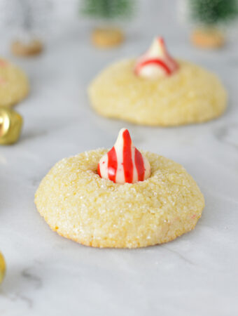 Peppermint Kiss Cookies with a red and white striped peppermint kiss on top of fluffy sanding sugar coated cookies. There are bells in the background, adding to the Christmas feel.