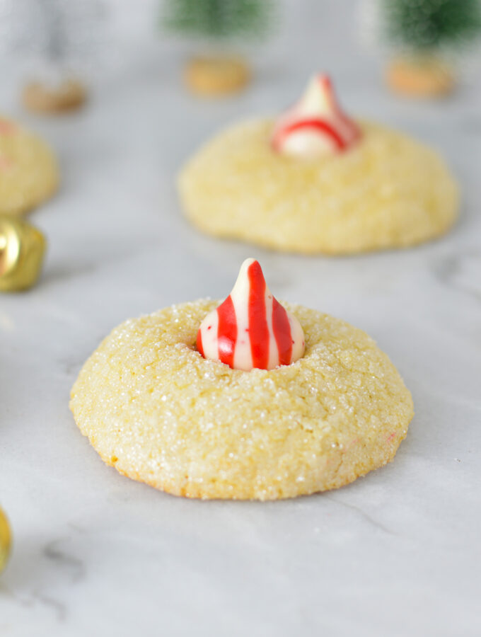 Peppermint Kiss Cookies with a red and white striped peppermint kiss on top of fluffy sanding sugar coated cookies. There are bells in the background, adding to the Christmas feel.