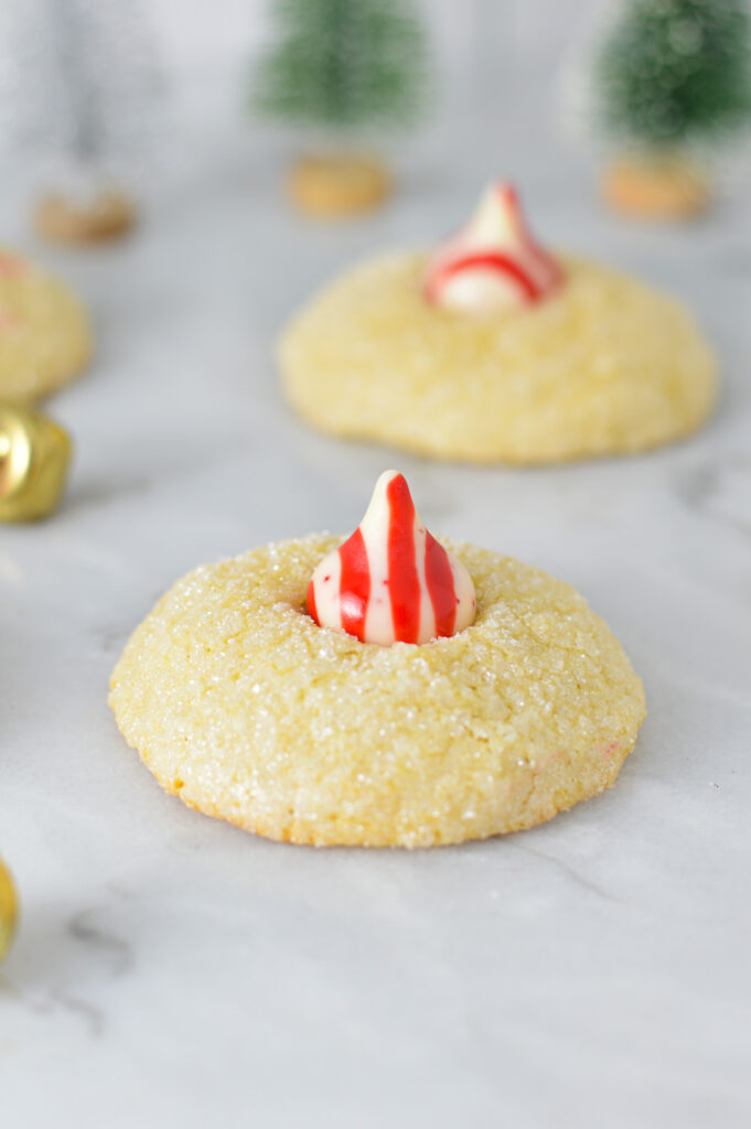 Peppermint Kiss Cookies with a red and white striped peppermint kiss on top of fluffy sanding sugar coated cookies. There are bells in the background, adding to the Christmas feel.