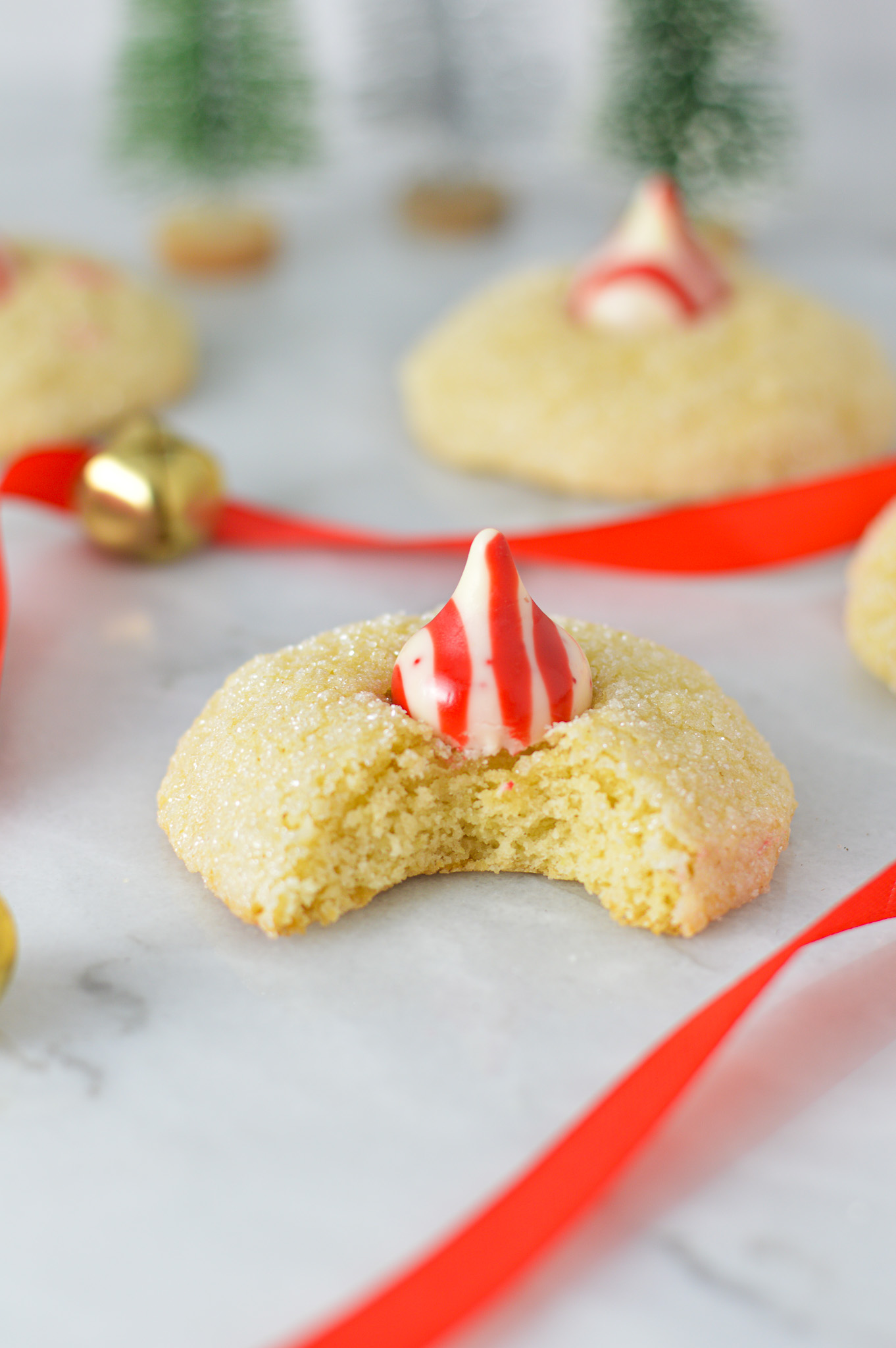 Peppermint Kiss Cookies with a bite taken out of one of the cookies revealing its soft and fluffy interior. Red ribbon and gold bells are in the background, adding to the Christmassy feel.