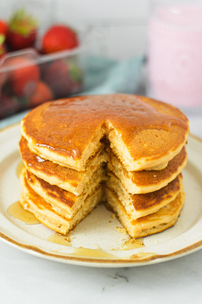 A stack of golden brown Strawberry Milk Pancakes, with a triangular slice taken out, showing the fluffy center of the pancakes.