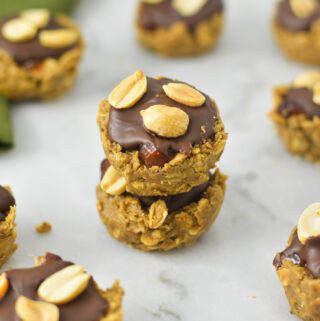 A stack of Mini Vegan Protein Snickers Cups on a granite surface. Each cup is topped with chocolate and dry roasted nuts.