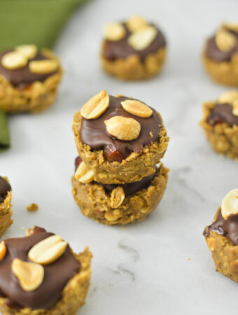 A stack of Mini Vegan Protein Snickers Cups on a granite surface. Each cup is topped with chocolate and dry roasted nuts.