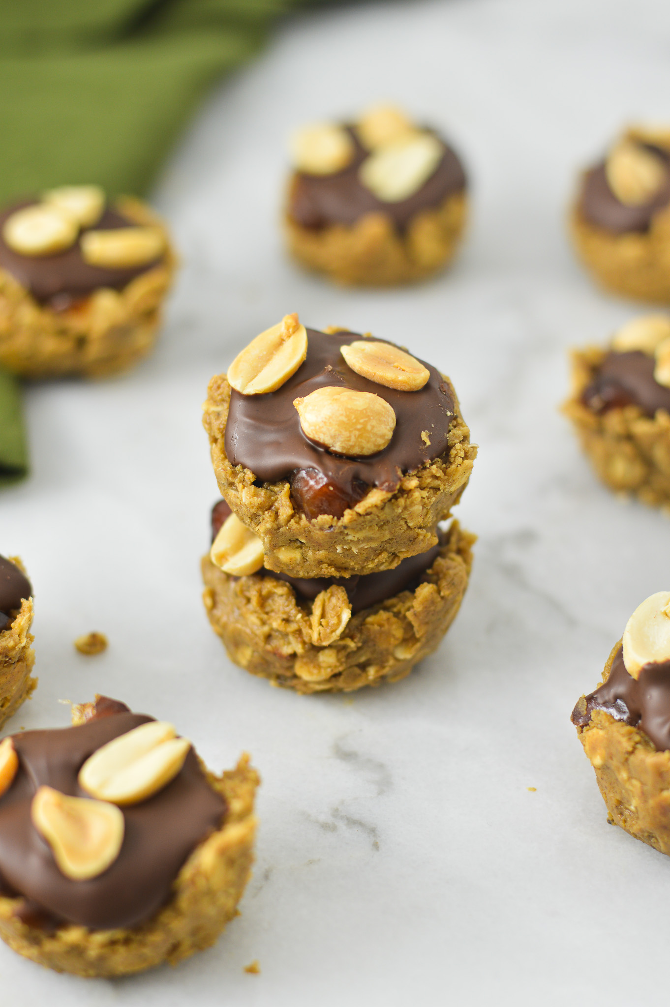 A stack of Mini Vegan Protein Snickers Cups on a granite surface. Each cup is topped with chocolate and dry roasted nuts.