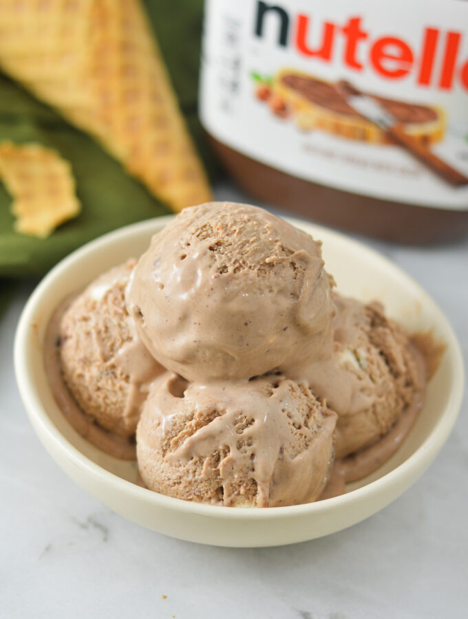 A shallow bowl filled with rich scoops of No Churn Nutella Ice Cream. A jar of Nutella and a broken up waffle cone are in the background.