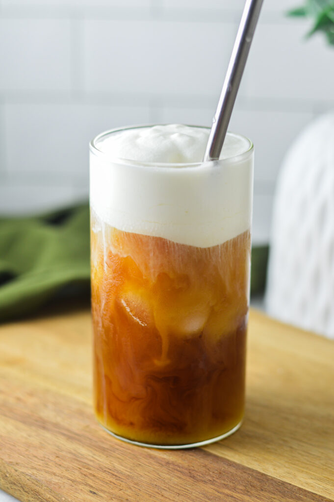 Iced coffee topped with Sweet Cream Cold Foam. A metal straw is sticking out of the tall glass cup, with swirls of creamy foam mixing into the cold coffee.