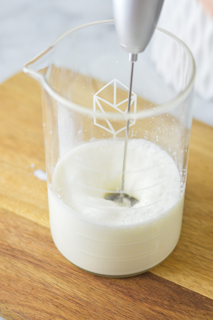 A handheld milk frother mixing some Sweet Cream Cold Foam in a tall glass jar.