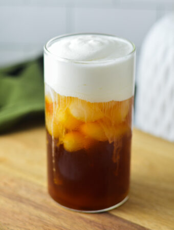 A tall glass of Sweet Cream Cold Foam on top of iced coffee. Condensation is visible on the glass, giving showing how cold this drink is.