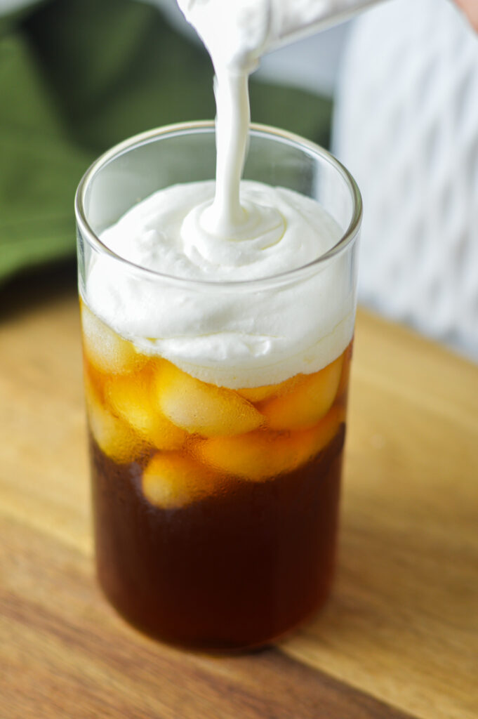 Sweet Cream Cold Foam being poured into a tall glass of iced coffee.