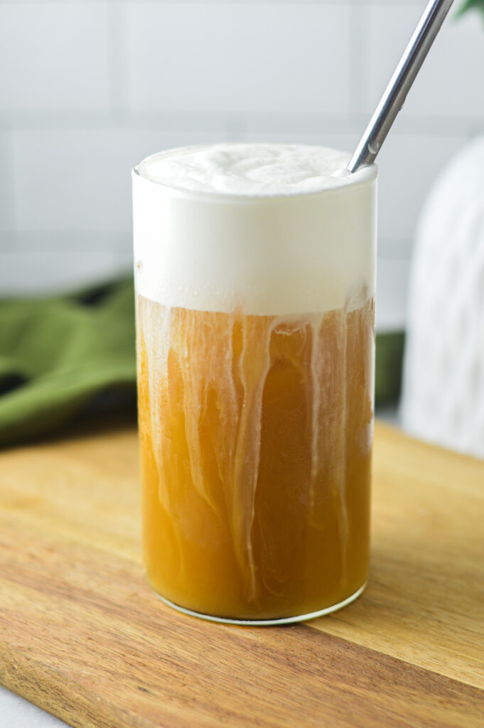 A tall glass filled with iced coffee, and topped Sweet Cream Cold Foam. A metal straw is coming out of the foam, and the coffee is already a golden brown color from the sweet cream mixing into it.