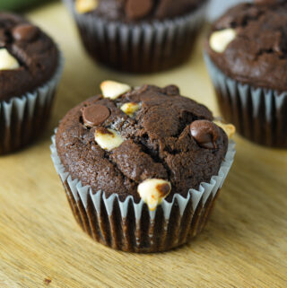 Fudgy Triple Chocolate Banana Muffins on a wooden board, with semi-sweet and white chocolate chips covering the tops of each muffin.