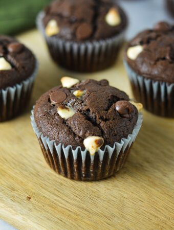 Fudgy Triple Chocolate Banana Muffins on a wooden board, with semi-sweet and white chocolate chips covering the tops of each muffin.