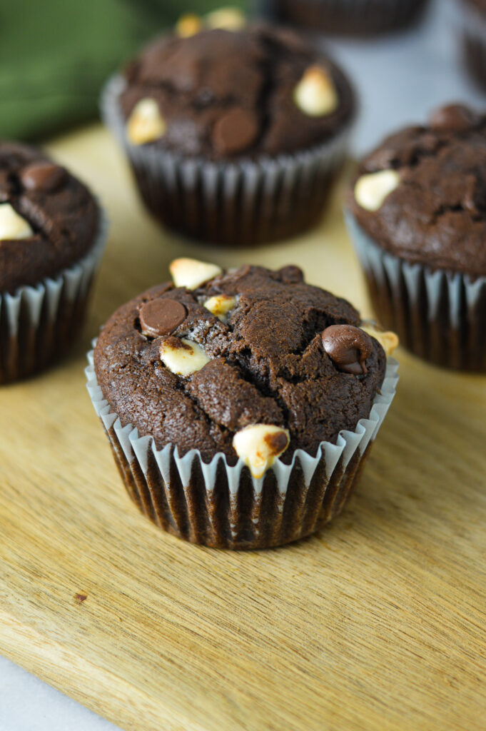 Fudgy Triple Chocolate Banana Muffins on a wooden board, with semi-sweet and white chocolate chips covering the tops of each muffin.