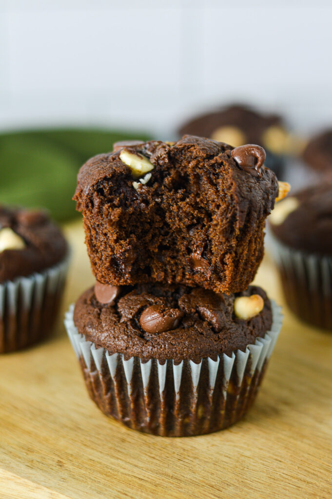 A stack of Triple Chocolate Banana Muffins on a wooden surface, with a bite taken out of the top muffin to show its fluffy texture.