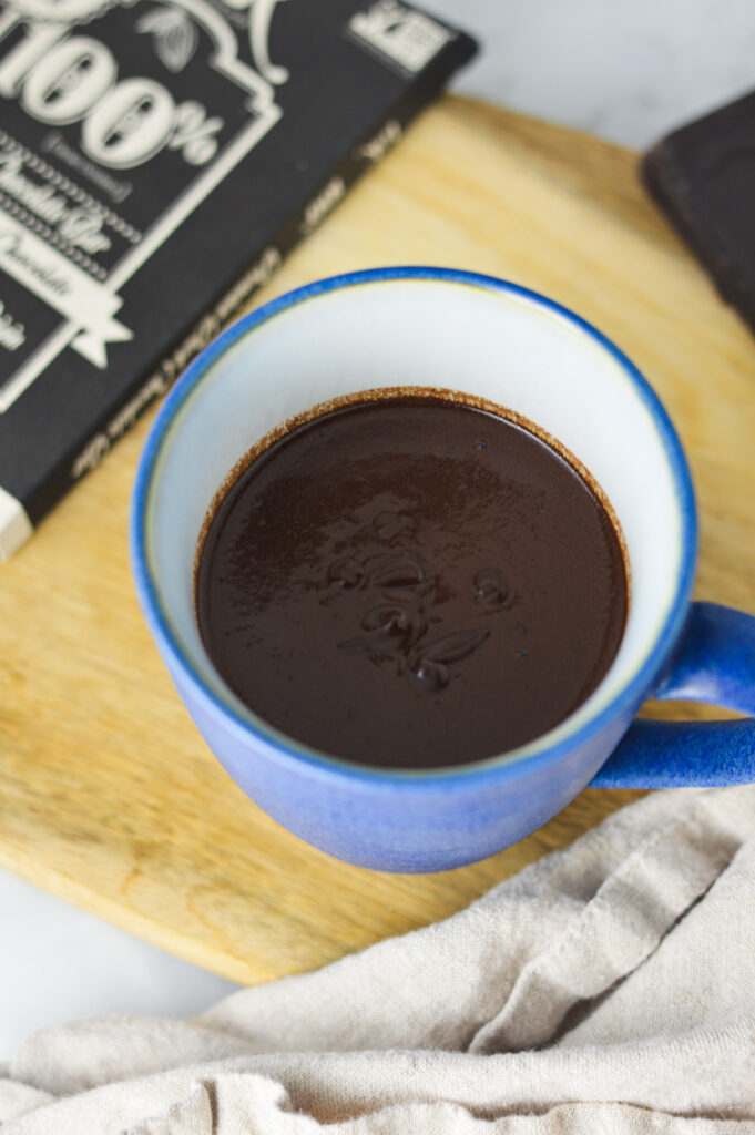 A mug full of dark, Rich Hot Chocolate, with a bar of 100% cocoa dark chocolate bar in the background.